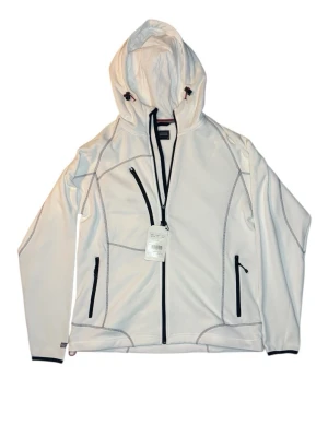 Vit vindjacka med huva från Jobman - Snygg vit vindjacka från Jobman med huva och svarta dragkedjor. Jackan har coola kontrastsömmar, tre fickor med dragkedja och justerbar huva med snörning. Perfekt för dig som gillar sportig stil och vill ha något som funkar när det blåser.