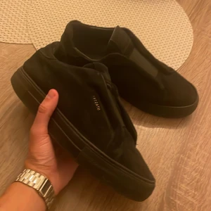 Svarta sneakers från Axel Arigato - Snygga svarta sneakers från Axel Arigato i mocka med diskret logga i guld på sidan. Skorna har en minimalistisk design, platt sula och rund tå. Perfekta för dig som gillar stilrena och trendiga skor. Pris kan  diskuteras!!