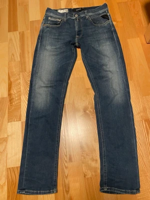 Replay Grover jeans blå, storlek 29 - Säljer ett par Replay Grover jeans i klassisk blå färg med snygga slitningar och raka ben. Jeansen har fem fickor, tydliga sömmar och coola detaljer på bakfickorna. Materialet är stretchigt jeans och passformen är slimfit, perfekt för en avslappnad stil.