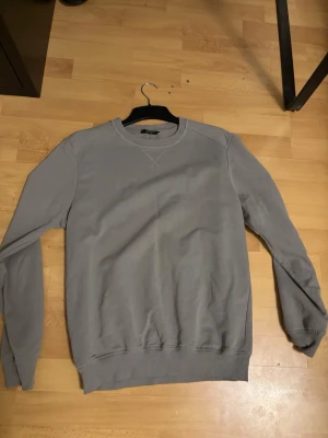 Grå sweatshirt med rund hals - En enkel grå sweatshirt från H&M med rund hals och ribbade muddar vid ärmslut och nederkant. Tröjan har lång ärm och är tillverkad i mjuk bomull, perfekt för chill dagar. Snygg att matcha med jeans eller joggers.