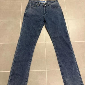 Blå jeans från Jacob Cohën, storlek 32 - Snygga blå jeans från Jacob Cohën i klassisk femficksmodell med silverfärgad knapp och dragkedja. Jeansen har ett stilrent, rakt snitt och är tillverkade i en mjuk bomullsblandning med 2% elastan för extra komfort. Diskret broderad J på myntfickan och coolt citat vid dragkedjan.