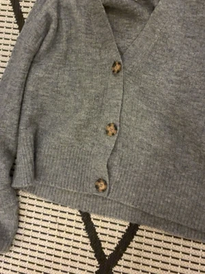 Grå kofta från 157 med knappar - Mysig grå kofta från 157 i storlek M passar även S lite oversize. Koftan har ribbade muddar, v-ringning och stora bruna knappar framtill. Perfekt att slänga på sig över en t-shirt eller linne när det är lite kyligare ute.