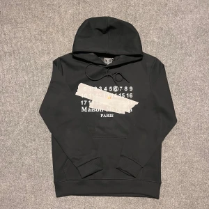 Maison Margiela Hoodie - storlek S - Maison Margiela-hoodie i mycket gott skick. Högkvalitativt material i klassiskt svart med logotypen framtill. Längd: 62 cm, bredd: 46 cm. Snabb frakt och snabba svar garanteras.