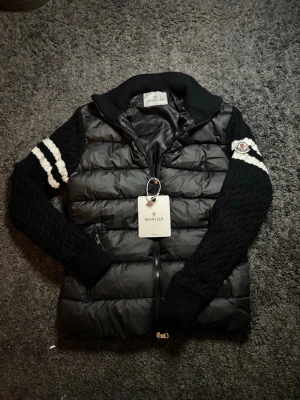 Hoodie  - Svart pufferjacka från Moncler med stickade ärmar och vita ränder på överarmarna. Jackan har dragkedja framtill, ribbad krage och Moncler-logga på ärmen. Kroppen är quiltad och tillverkad i blankt syntetmaterial, medan ärmarna är stickade med kabelmönster.