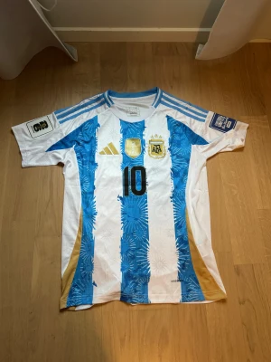 Argentina Messi tröja VM 2022 - Säljer en officiell Argentina landslagströja från VM 2022 med nummer 10 och Messi på ryggen. Tröjan har blå och vita ränder, guldiga detaljer och broderade märken. Materialet är lätt polyester och har snygga tryck på ärmarna. Perfekt för fotbollsfans!
