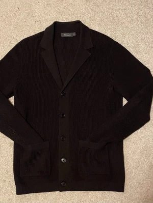 Cardigan från Dressmann - Mörkbrun ribbstickad cardigan jacka från Dressmann med klassisk krage och knäppning framtill. Jackan har två framfickor och är långärmad, perfekt för lager-på-lager. Materialet är stickat och ger en skön känsla. Snygg och enkel att matcha med olika outfits.