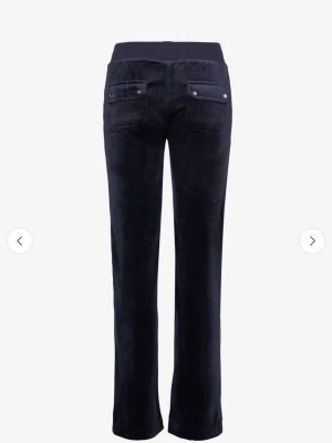 Juicy Couture - Mörkblå mjukisbyxor i mjuk sammet med raka ben och bred resårmidja med snörning. Byxorna har fickor fram och bak med snygga knappdetaljer. Perfekta för chill dagar eller när du vill vara extra bekväm. Helt ny skick