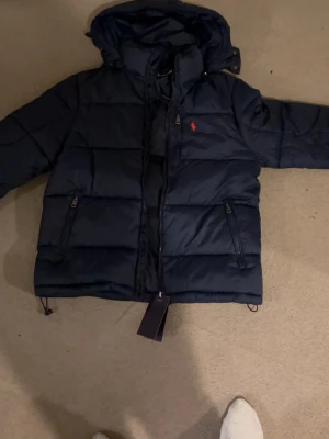 Mörkblå dunjacka från Polo Ralph Lauren - Snygg mörkblå dunjacka från Polo Ralph Lauren med röd logga på bröstet. Jackan har huva, dragkedja framtill och två sidofickor med dragkedja. Perfekt för kalla dagar och har en klassisk pufferstil.