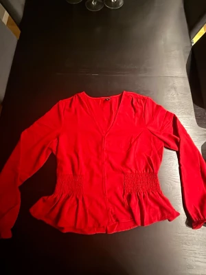Röd blus med smock från Vero Moda - Snygg röd blus från Vero Moda med v-ringning och smockdetaljer i midjan. Blusen har långa ärmar och en lätt utsvängd nederkant som ger en feminin siluett. Tillverkad i mjukt tyg som känns skönt mot huden.