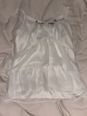 Vit linnetopp med pärldetaljer - Söt vit linnetopp med tunna axelband och volangdetaljer. Toppen har dekorativa pärlor och små stenar längs ringningen som ger en extra touch. Tillverkad i ett lätt, skirt material som känns luftigt och somrigt.