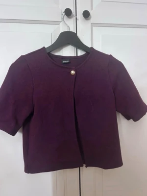 Lila kort kofta från Gina Tricot - Kort lila kofta från Gina Tricot med rund halsringning och korta ärmar. Koftan har en dekorativ knapp framtill i guld och är gjord i ett mjukt tyg. Perfekt att slänga över en topp för en snygg lager-på-lager-look.