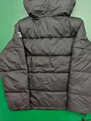 Svart dunjacka från Burberry - Snygg svart dunjacka från Burberry med huva och tydlig logga på ärmen. Jackan har quiltad design, dragkedja och knappar framtill samt sidofickor. Perfekt för kalla dagar när du vill hålla stilen.