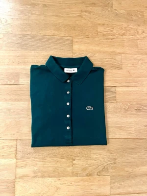 Mörkgrön pikétröja från Lacoste - Snygg mörkgrön pikétröja i slim fit, från Lacoste. Tröjan har vita knappar framtill och den fina krokodilloggan broderad på bröstet. Pikétröjan är i storlek 38, som motsvarar 158. Skriv till mig vid minsta fundering!