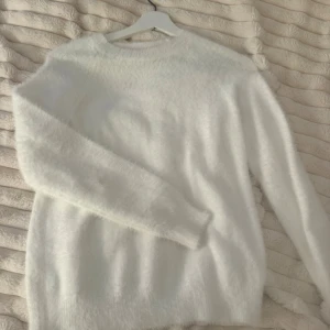 Vit fluffig stickad tröja - Mysig vit stickad tröja med fluffig yta och rund hals. Tröjan har långa ärmar och en avslappnad passform, perfekt för kalla dagar. Materialet känns mjukt och gosigt mot huden. Enkel men stilren design som passar till det mesta.
