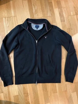 Svart ziptröja från GANT - Klassisk svart ziptröja från GANT med dragkedja framtill och två fickor. Tröjan har ståkrage med diskreta rödvita detaljer på insidan och GANT-logga broderad på bröstet. Perfekt till jeans eller över en t-shirt för en avslappnad look.