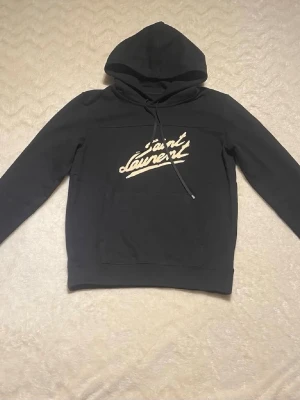 Svart hoodie från Saint Laurent - Snygg svart hoodie från Saint Laurent med vit logga tryckt på bröstet. Klassisk modell med huva och dragsnören samt ribbade muddar vid ärmar och midja. Perfekt för en avslappnad och trendig stil. Tillverkad i mjukt material för extra komfort.