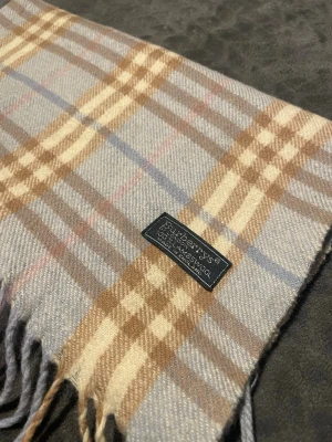 Rutig halsduk från Burberrys i ull - Klassisk halsduk från Burberrys i mjuk ull med beige, brunt, blått och grått rutmönster. Halsduken har fransar i ändarna och den ikoniska Burberrys-loggan på en tygpatch. Perfekt accessoar för att lyfta din outfit.