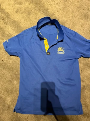 Blå pikétröja från Burberry - Snygg blå pikétröja från Burberry med gul logga och text på bröstet. Kragen har en gul insida som ger en cool kontrast. Klassisk pikémodell med kort ärm och knappslå framtill. Perfekt för en stilren och sportig look.