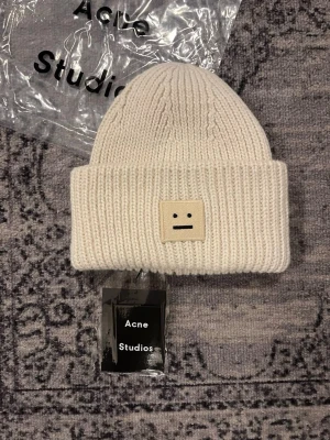 mössa Acne Studios - Säljer en stilren vit ribbstickad mössa från Acne Studios med uppvikt kant och det klassiska ansiktslogotypen framtill. Mössan är gjord i mjukt och varmt material, perfekt för kalla dagar. Enkelt och trendigt plagg som passar till det mesta.