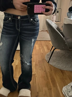 Mörkblå bootcut jeans med slitningar - Säljer ett par mörkblå bootcut jeans med vita kontrastsömmar och slitna detaljer framtill. Jeansen har broderad bakficka och klassisk femficksmodell. Materialet är jeans i bomull och passformen är normal med låg midja.  På bild 4 ser ni att kanterna har ett litet jak. Klippte båda lite men syns knappast då man kan ha en tröja över. 