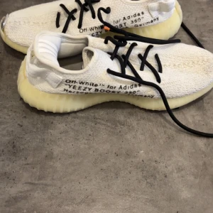 Vita Adidas Yeezy Boost sneakers - Säljer ett par vita Adidas Yeezy Boost sneakers med svart snörning och textdetaljer på sidan. Skorna har en ribbad, ljusgul sula och är tillverkade i ett stickat syntetmaterial för en modern och sportig look.
