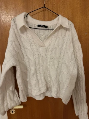 Vit kabelstickad tröja med krage - Säljer en vit kabelstickad tröja från Bikbok med klassisk krage och v-ringning. Tröjan har långa ärmar och en avslappnad passform, perfekt för lager på lager. Mjuk och skön stickning som ger en trendig look.
