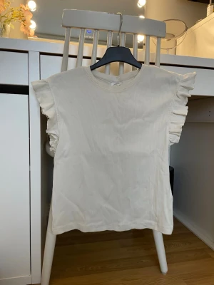 Vit ribbad topp med volangärmar - Supersöt vit ribbad topp med volangdetaljer på ärmarna. T-shirten har rund halsringning och är tillverkad i ett mjukt bomullsmaterial. Perfekt för dig som gillar en feminin och trendig stil.