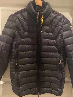 Svart dunjacka från Parajumpers - Snygg svart dunjacka från Parajumpers med gul detalj vid halsen och quiltad design. Jackan har hög krage, dragkedja framtill och två sidofickor med dragkedja. Perfekt för kalla dagar och har en klassisk pufferlook. Jackan är liten i storlek, liknar mer en M