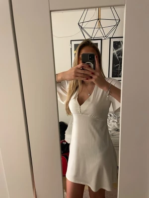 Superfin vit kort klänning🤍 - Snygg vit kortklänning med v-ringning och knappar framtill. Klänningen har korta ärmar och är gjord i ett ribbat material som ger en bekväm och snygg look. Perfekt till student, midsommar eller skolavslutning.🌸🤍