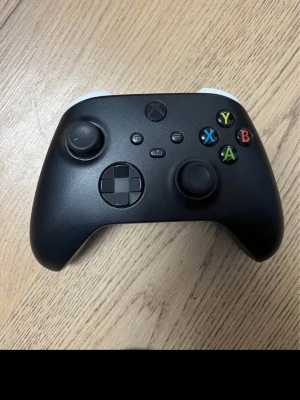 Svart Xbox handkontroll - Snygg svart Xbox Series-handkontroll med klassisk layout, färgade knappar (A, B, X, Y) och greppvänlig form. Tillverkad i slitstark plast med vita detaljer på baksidan och USB-C-port för laddning eller anslutning.