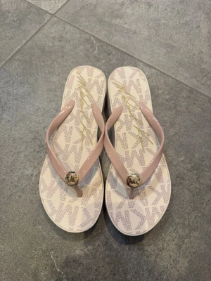 Beige sandaler från Michael Kors - Snygga beige sandaler från Michael Kors med plattformsula och ljusrosa remmar i syntet. Dekorerade med guldfärgad MK-logga sopm detalj framtill. Mönstrad sula med Michael Kors-loggor i beige och guld. Perfekta för sommaren!