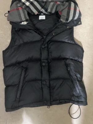 Svart dunväst med huva från Burberry - Svart puffig dunväst från Burberry med avtagbar huva som har klassiskt rutigt foder i grått, vitt och rött. Västen har dragkedja och knappar framtill samt två fickor med dragkedja. Perfekt för lager på lager och en riktigt clean look.