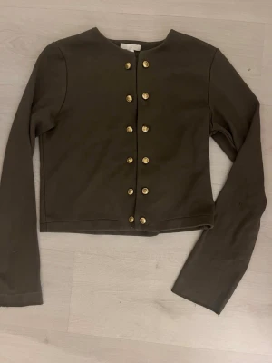 Mörkgrön kofta med guldknappar H&M - Stilren mörkgrön kofta från H&M med raka linjer och dekorativa guldknappar framtill. Kortare modell med rund halsringning och långa ärmar. Perfekt för att lyfta en enkel outfit med lite extra detaljer. Dock en liten slitning vid handleden