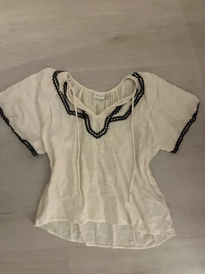 Vit blus med svarta detaljer från Vila - Söt vit blus från Vila med korta ärmar och svarta vågiga band längs halsringningen och ärmsluten. Blusen har knytband vid halsen och är tillverkad i ett lätt, luftigt tyg som ger en avslappnad känsla.