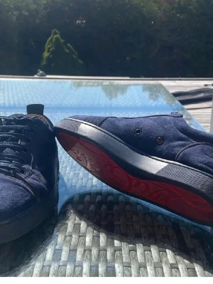 Louboutins strlk 42 - Snygga mörkblå sneakers i mocka med svart snörning och unik röd sula. Skorna har låg profil och rund tå, samt diskreta detaljer på hälen. Perfekta för dig som vill ha något stilrent men ändå sticker ut. Slitna innuti, men inget jag stört mig på,kan fixa fler bilder