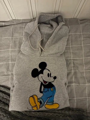Grå Levi’s hoodie med Mickey Mouse-tryck - Säljer en grå Levi’s hoodie med stort färgglatt tryck av Mickey Mouse på framsidan. Hoodien har huva och en klassisk känguruficka. Perfekt för dig som gillar retro och tecknade figurer. Materialet är mjukt och skönt bomullstyg. Den är otroligt skön och mjuk. Näst intill ny skick har använts ett väldigt få antal gånger.