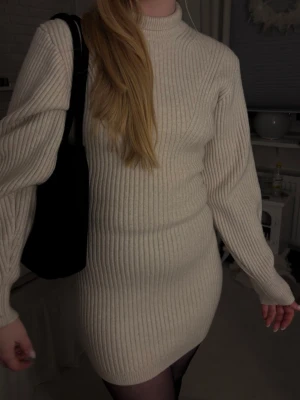 Ribbad beige stickad kortklänning - Säljer en ribbad beige stickad klänning med lång ärm och hög hals. Klänningen är kort och har en kroppsnära passform som framhäver figuren. Perfekt att styla med boots och strumpbyxor för en trendig look.