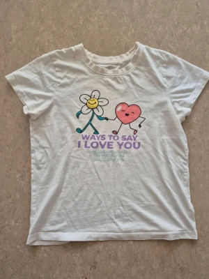 T-shirt - Vit t-shirt i bomull med ett gulligt tryck av en blomma och ett hjärta som håller hand. Texten 'Ways to say I love you' i lila och blått framtill. Klassisk rund hals och korta ärmar. Perfekt för dig som gillar söta motiv och enkel stil. Finns rosa fläckar.