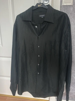 Svart randig skjorta från Atlant - Snygg svart skjorta från Atlant med tunna vertikala ränder och klassisk krage. Skjortan har lång ärm, knappar framtill och en bröstficka. Perfekt för en stilren look och passar till många olika outfits.