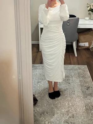 Vit lång studentklänning med rynkade detaljer - Elegant vit långklänning med långa ärmar och ryn sopplad effekt längs sidan som ger en smickrande siluett. Klänningen har en bred, båtringad urringning och är tillverkad i ett mjukt, stretchigt material som formar sig efter kroppen.