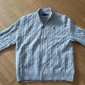 Grå kabelstickad kofta från Polo Ralph Lauren - Snygg grå kabelstickad kofta från Polo Ralph Lauren med dragkedja framtill och två fickor. Klassisk design med ribbade muddar och diskret logga broderad på bröstet. Perfekt för lager på lager och en stilren look.