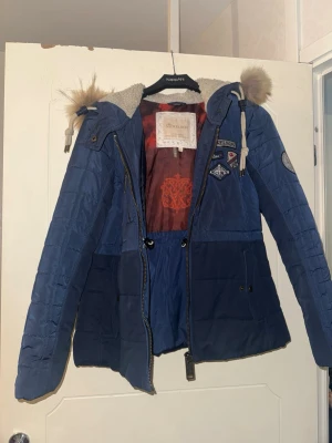 Blå dunjacka från Nickelson - Snygg blå dunjacka från Nickelson med äkta päls på huvan och coola patchdetaljer på bröstet. Jackan har dragkedja, snörning i midjan och mysigt foder. Perfekt för kalla dagar och ger en sportig vibe.