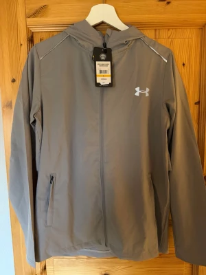 Grå vindjacka från Under Armour - Helt ny Windbreaker från Under Armour. Storlek L och alla tags e kvar. Tveka inte vid att höra av er! 