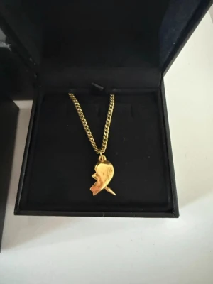 Guldhalsband Maria Nilsdotter - Unikt halsband i guld från Maria Nilsdotter med en cool, abstrakt berlock i form av ett ansikte. Kedjan är grov och har en exklusiv känsla. Perfekt för dig som vill ha något personligt och trendigt i smyckesamlingen.