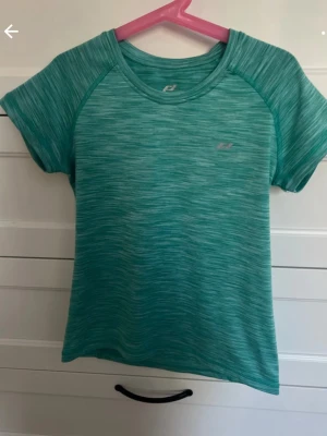 Grön träningströja Pro Touch - Säljer en grönmelerad träningströja från Pro Touch med korta ärmar och rund hals. Tillverkad i funktionsmaterial med DRY PLUS-teknologi som transporterar bort fukt, perfekt för träning och sportaktiviteter.