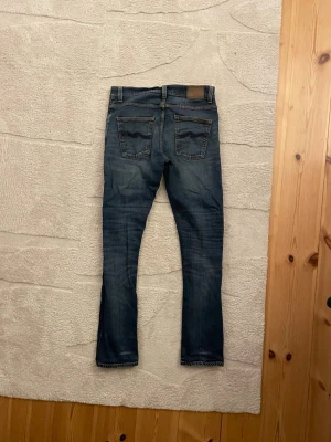Nudie Jeans Grim Tim - Snygga mörkblå jeans från Nudie med klassisk femficksdesign och diskreta slitningar framtill. Jeansen har smal passform och raka ben, tillverkade i mjuk bomullsdenim. Perfekta för dig som gillar en stilren och modern look. Modellen är Grim Tim och sitter som Slim fit. Priset kan diskuteras
