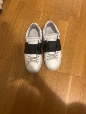 Valentino Open sneakers vit/svart - Snygga Valentino Open sneakers i vitt skinn med bred svart rem över vristen. Klassisk låg modell med rund tå och vita snören. Perfekta för dig som gillar stilrena och exklusiva sneakers med en twist.