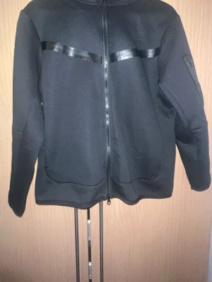 Svart Nike tech dress -  en svart Nike tech fleece dress. Den har huva och fickor framtill samt en extra ficka på ena ärmen. Materialet är mjukt och stretchigt, perfekt för en chill och stilren look.