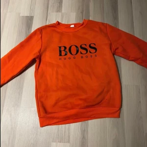 Orange sweatshirt från Hugo Boss - Säljer en snygg orange sweatshirt från Hugo Boss med stor svart BOSS-logga på bröstet. Tröjan har rund halsringning och långa ärmar, perfekt för dig som vill sticka ut med en färgstark och trendig look.