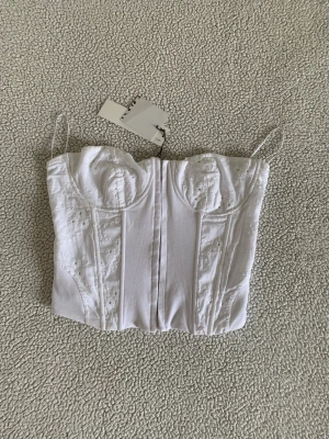 ZARA vit korsett med hålbroderier - Snygg vit korsett med broderade spetsdetaljer. Modellen har markerade kupor och dekorativa sömmar framtill för en feminin siluett. Perfekt att styla till jeans eller kjol för en trendig look. Ny med prislapp. Storlek S eller M om jag inte minns fel. 